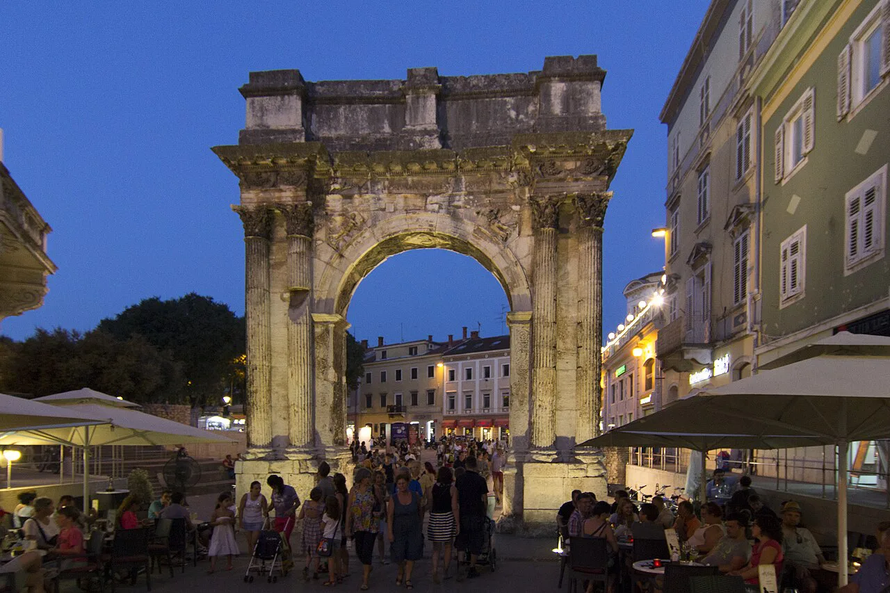 Café tables by the Arch of the Sergii, Pula, at dusk.